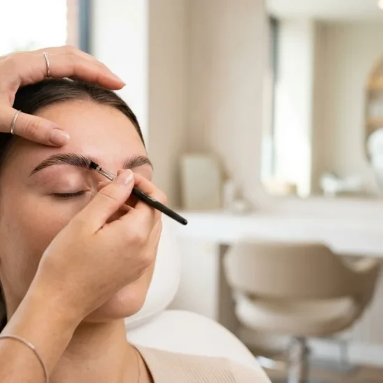 What Are HD Brows? A close-up photograph focuses on a client’s eyebrow as a beauty professional applies dark brow tint with a fine-tipped brush. The client is relaxed with eyes closed, leaning back in a plush white salon chair. The stylist’s hands, adorned with delicate silver rings and a bracelet, are precise as they apply the product. The modern salon background is softly blurred with warm, neutral tones.